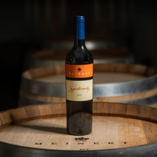 A bottle of Meinert Synchronicity standing on an oak barrel